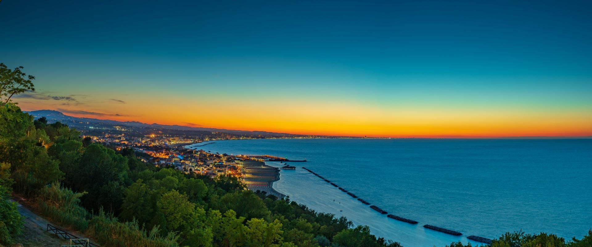 Tramonto sulla costa con città illuminata e mare calmo.
