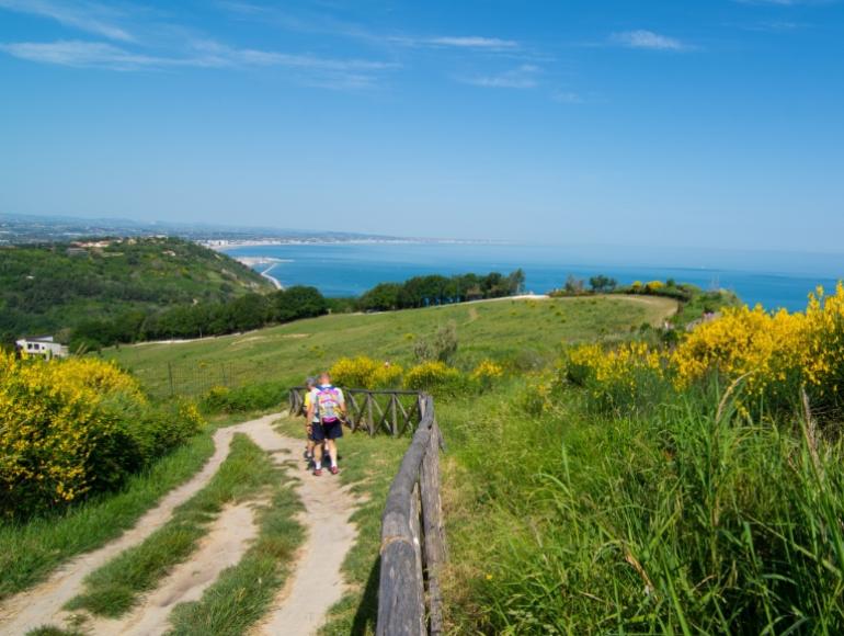 Coppia cammina su sentiero panoramico con vista mare e colline verdi.