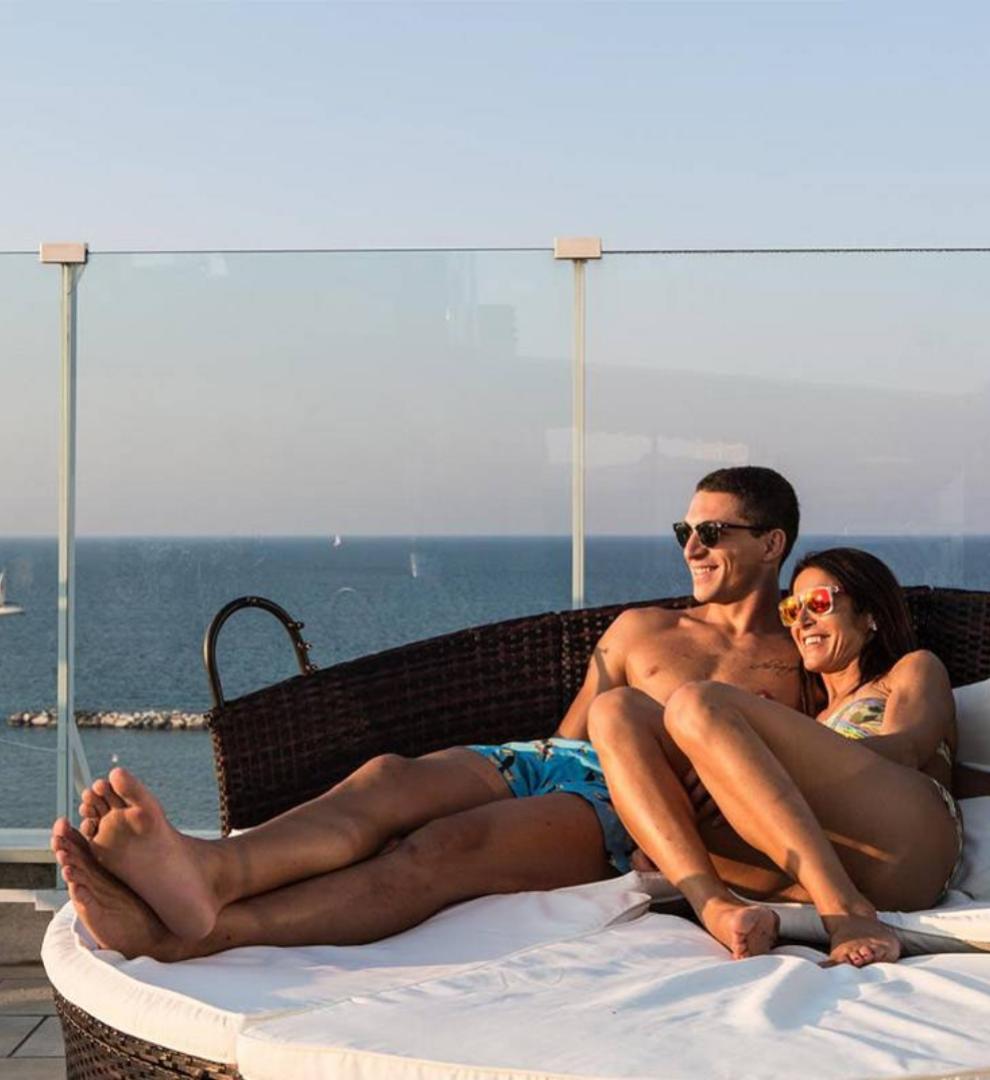 Coppia rilassata su terrazza con vista mare, seduta su un lettino.